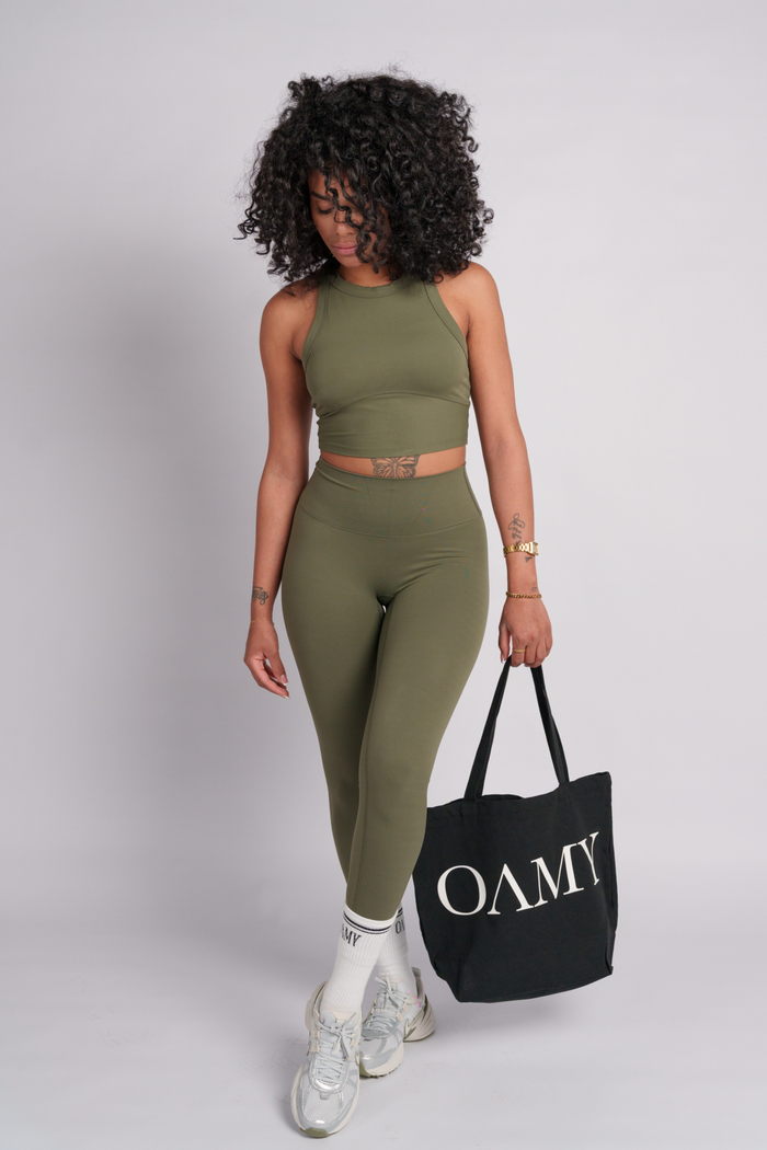 Woman in olive green crop top and leggings holding a black tote bag with 'OAMY' branding.